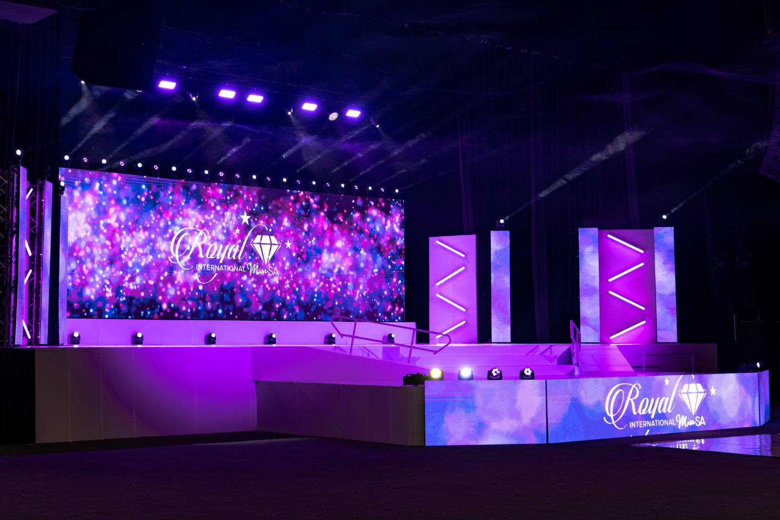 A stage from the side: LED wall, lit towers and moving heads on the deck, the hall dark above